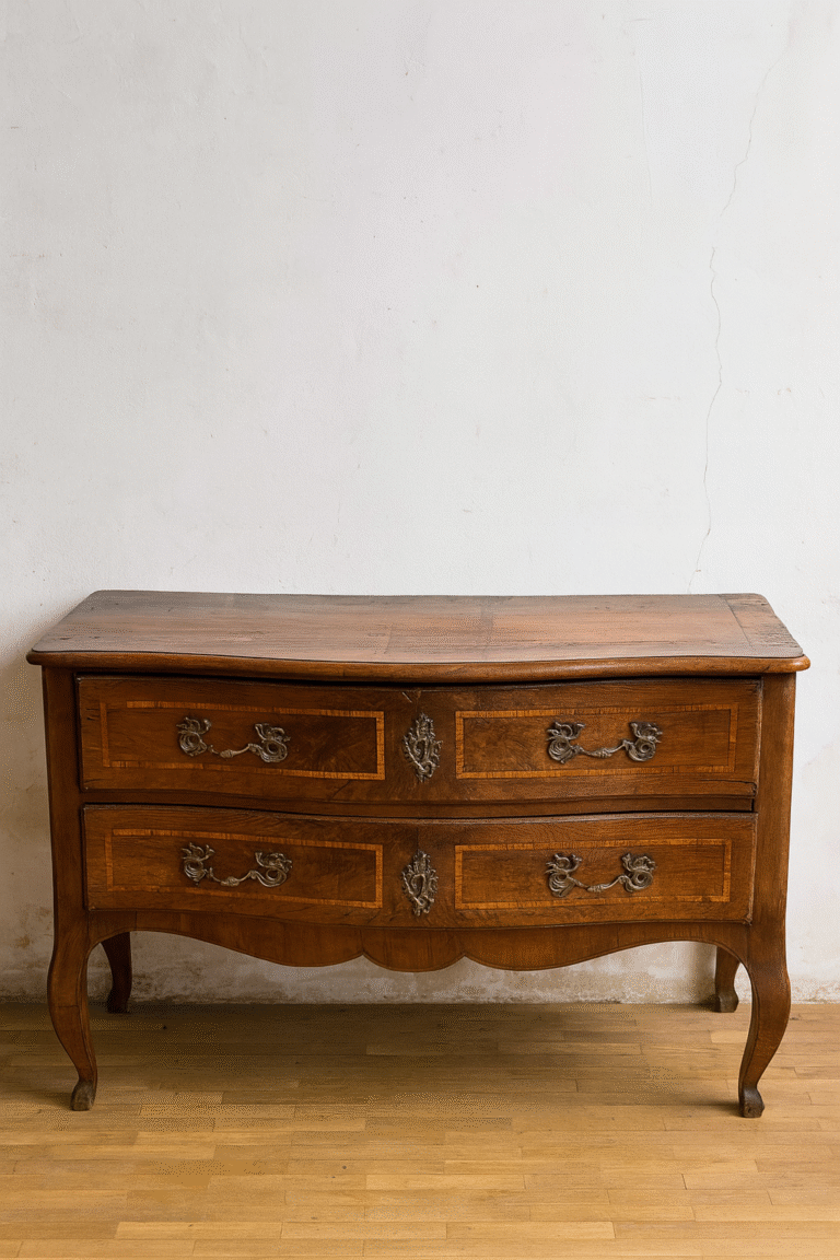 Cómoda antigua de madera con dos amplios cajones decorados con marquetería enmarcada y tiradores metálicos ornamentales. El frente ligeramente curvo y las patas cabriolé suavemente arqueadas aportan elegancia y movimiento. Ideal como mueble auxiliar en dormitorios o recibidores con encanto clásico.